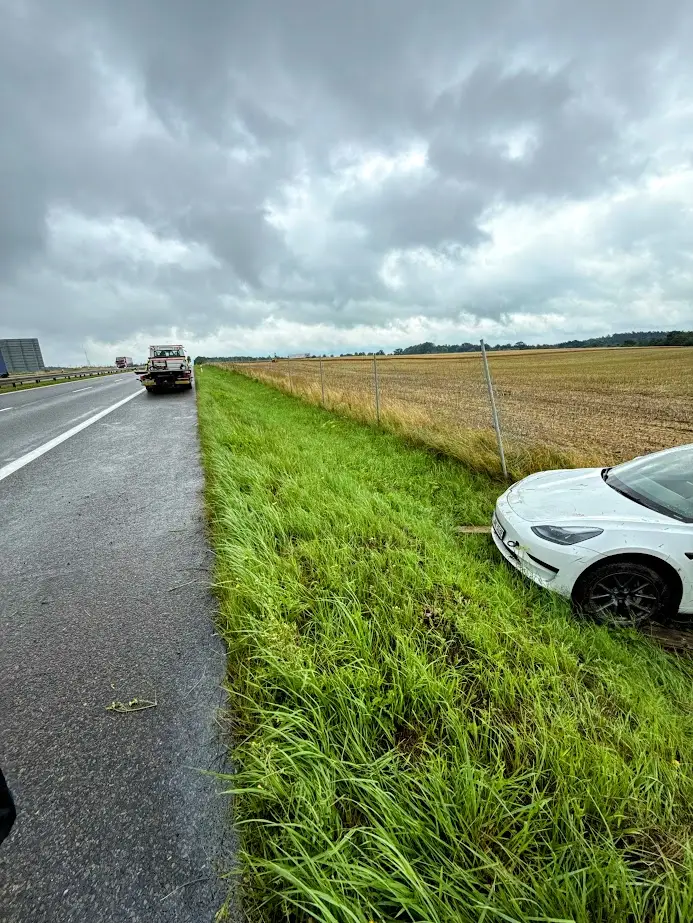 Biały samochód osobowy znajduje się w rowie na trawiastym poboczu przy drodze. W oddali na pasie awaryjnym widać tył zaparkowanej pomocy drogowej pod zachmurzonym niebem.