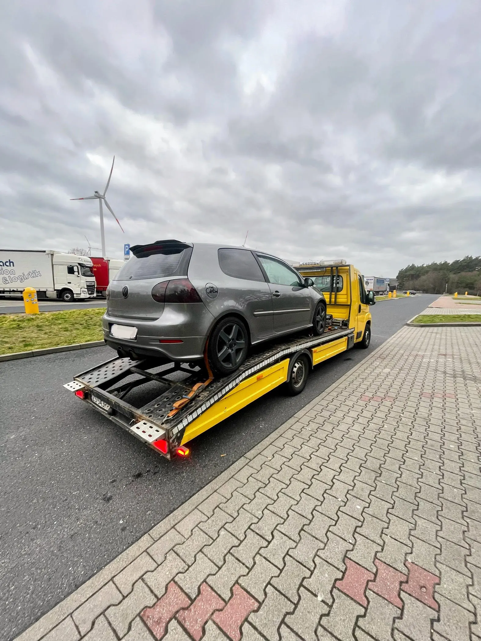 Srebrny Volkswagen Golf hatchback z zerwanym paskiem rozrządu, bezpiecznie zabezpieczony pasami transportowymi na platformie żółtej autolawety Fiat Ducato firmy KM Transport Szczecin. Samochód stoi na parkingu MOP przy drodze ekspresowej S3, gotowy do transportu do warsztatu.
